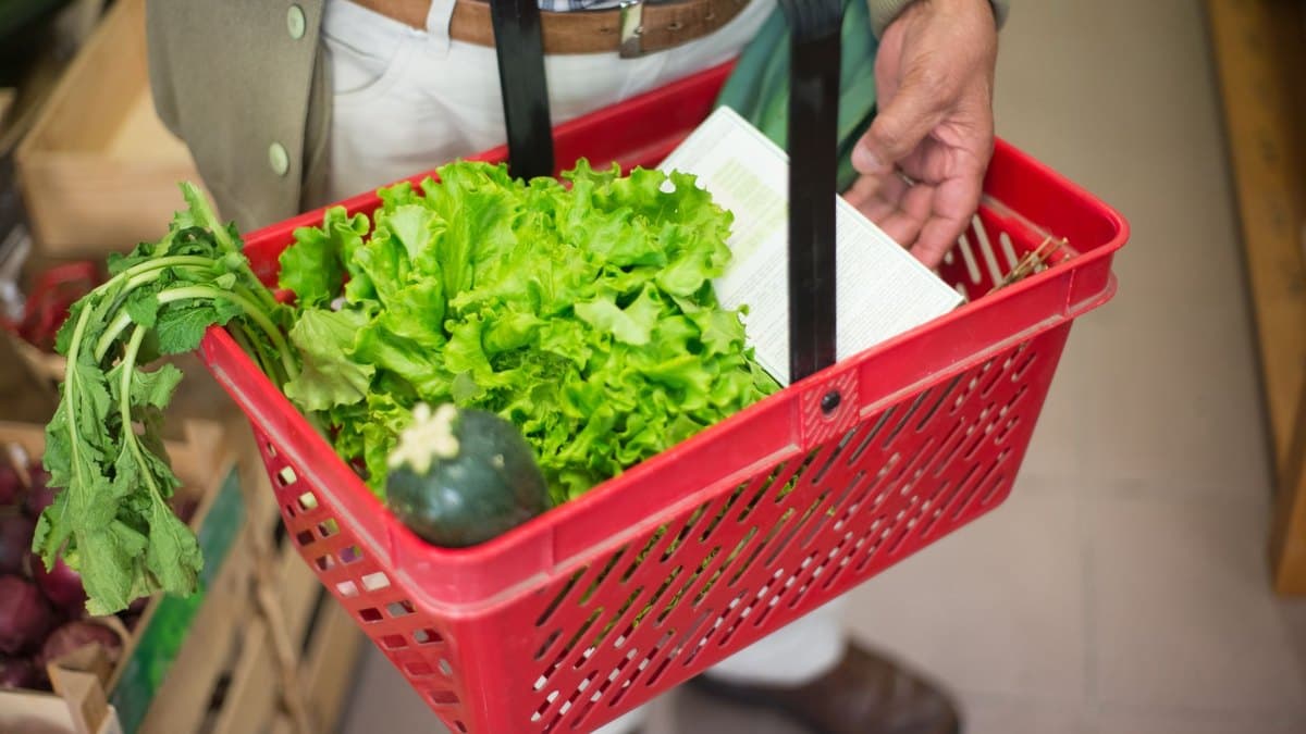 A shopper carrying a supermarket basket of fresh vegetables and greens